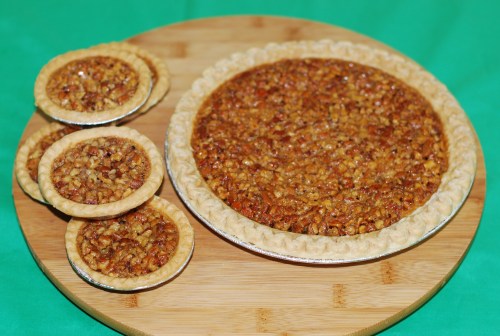 A large pecan pie and three smaller pecan tarts on a wooden cutting board against a green background.