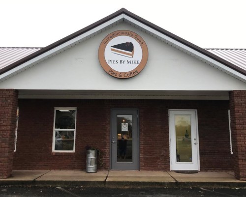 Exterior of "Pies By Mike" shop featuring a sign and brick facade, with a door on the right and a barrel outside.