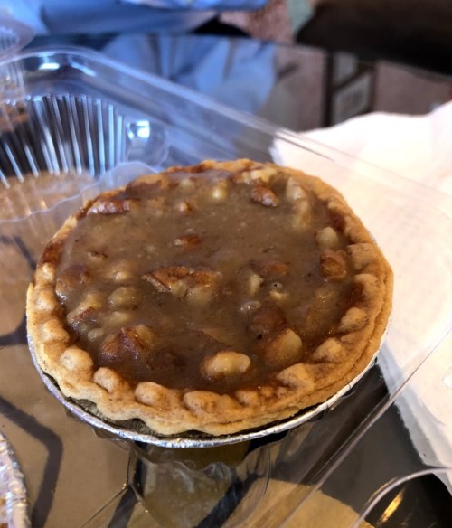 A small pie with a golden crust filled with a thick, brown filling and bits of nuts, placed on a clear container.