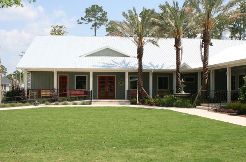 A modern building with a metal roof, red doors, and palm trees, set in a green lawn under a blue sky.