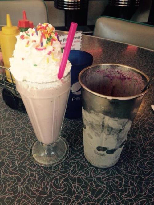 A tall milkshake topped with whipped cream and sprinkles, next to a metal cup with remnants of the shake.