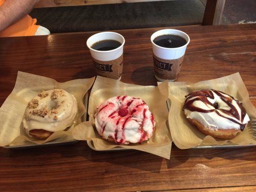 Three donuts on a tray: one with nuts, one with red icing, and one with chocolate drizzle, alongside two cups of coffee.