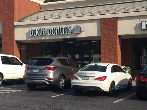 Exterior of Duck Donuts shop with two parked cars in front, featuring a brick facade and a sign above the entrance.