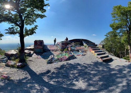 Two people sit on a colorful, graffiti-covered rock formation with steps, surrounded by trees and a clear blue sky.