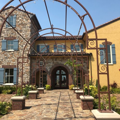 A stone and yellow building with an arched trellis entrance, surrounded by greenery and a stone pathway.