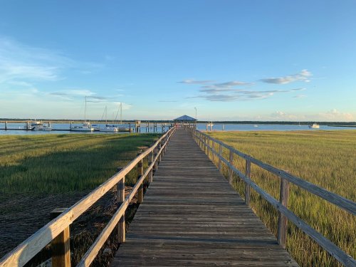 A wooden pier extends over a calm waterway, surrounded by green marsh grass and boats in the distance under a clear sky.