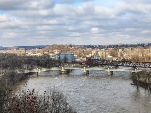 A view of a river with a bridge, surrounded by trees and buildings under a cloudy sky.