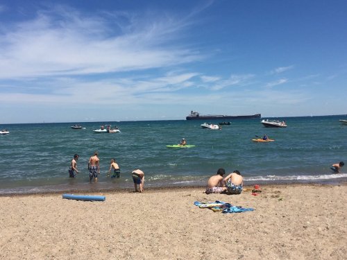 People enjoying a sunny day at the beach, swimming and boating in the water with a cargo ship in the background.
