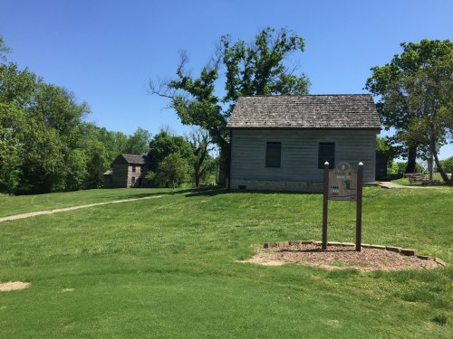 A grassy area with two historic buildings and a sign, under a clear blue sky with a few trees in the background.