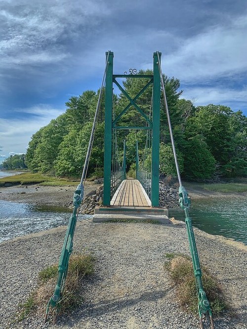 A green suspension bridge spans a calm waterway, surrounded by lush trees and a cloudy blue sky.