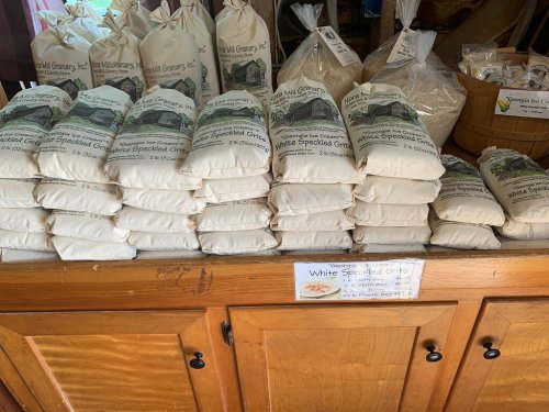 Bags of white speckled grits stacked on a wooden counter, with a sign displaying the product name.