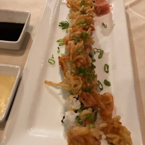 A plate of sushi topped with crispy tempura, garnished with green onions, served with dipping sauces on the side.