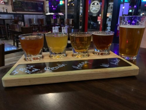 A wooden flight board with five small glasses of different colored beers and a pint glass on a table in a bar.