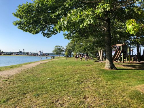A sunny park by a river with trees, a playground, and people walking along a grassy path.