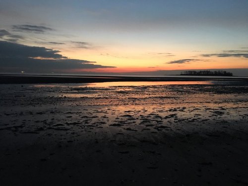 A serene sunset over a calm beach, with reflections on the wet sand and a distant island silhouette.