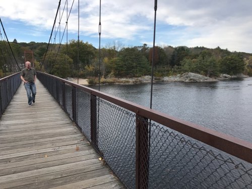 A person walks on a wooden suspension bridge over a calm river, surrounded by trees and rocky shores.