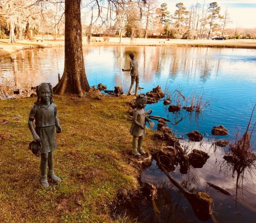 Three bronze statues of children stand by a serene pond, surrounded by trees and reflecting water.