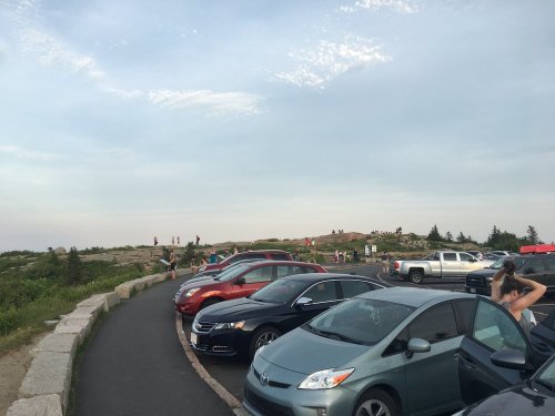A parking area with various cars, people walking on a rocky hill in the background, and a cloudy sky above.