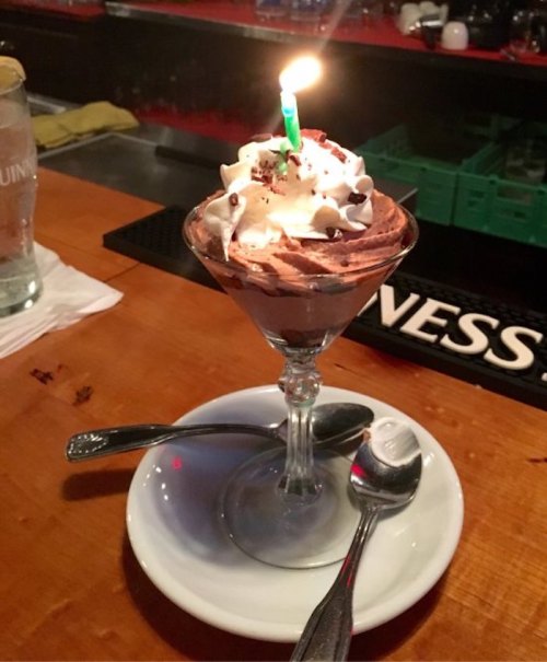 A dessert in a glass with chocolate mousse, whipped cream, chocolate sprinkles, and a lit green candle on top.