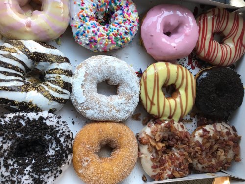 A variety of colorful donuts in a box, featuring sprinkles, frosting, and toppings like bacon and chocolate.