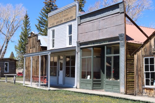 Historic wooden buildings in a ghost town, featuring a bakery sign and a green storefront, surrounded by trees.