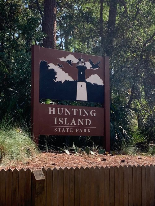Sign for Hunting Island State Park featuring a lighthouse and birds, surrounded by trees and greenery.