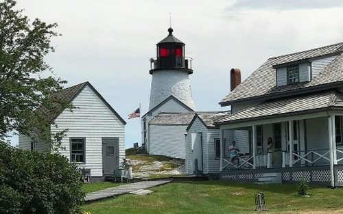 A white lighthouse with a red top stands near a house, surrounded by green grass and trees under a cloudy sky.
