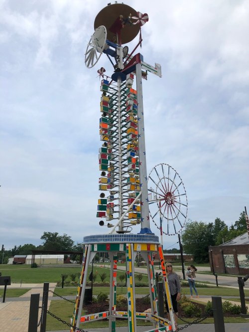 A colorful, tall windmill sculpture with spinning elements and a ferris wheel in the background, set in a park.