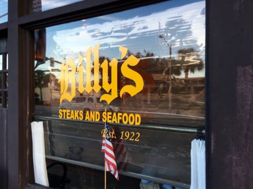Sign on a restaurant window reading "Billy's Steaks and Seafood, Est. 1922," with an American flag visible.