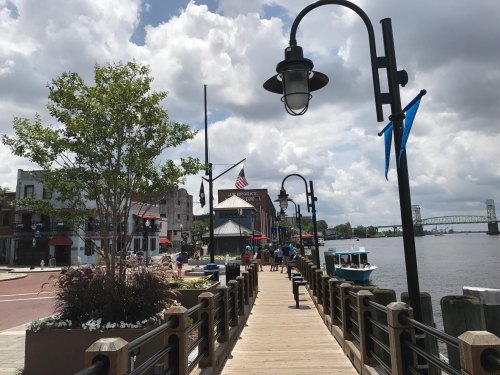 A scenic waterfront boardwalk with shops, boats, and a bridge in the background under a partly cloudy sky.