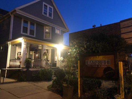 A cozy restaurant exterior at dusk, featuring a welcoming porch and a sign reading "Batuuli: The Flavor of Basil."