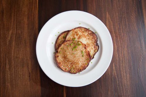 Three golden-brown pancakes topped with chopped green herbs, served on a white plate against a wooden background.