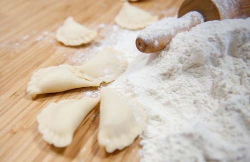 A wooden rolling pin rests on a surface with flour and uncooked pasta shapes scattered around.