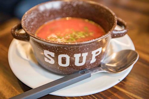 A bowl labeled "SOUP" filled with red soup, garnished with green herbs, on a white plate with a spoon beside it.