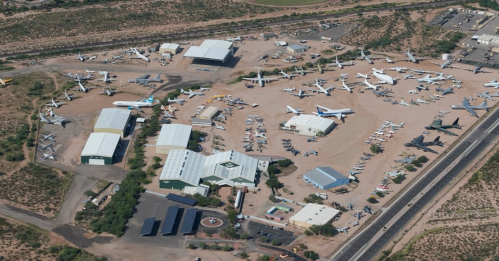 Aerial view of an aircraft boneyard with numerous planes parked on a dusty lot surrounded by buildings and roads.
