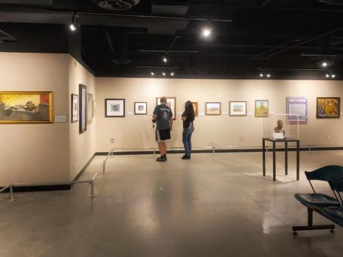 Two visitors observe artwork in a gallery, surrounded by framed pieces on the walls and a sculpture on display.