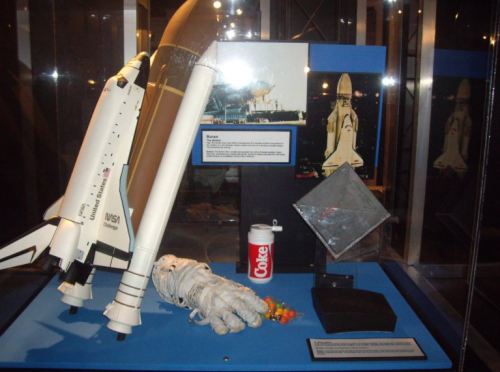 Display case featuring a model space shuttle, astronaut gloves, a Coke can, and a reflective panel.