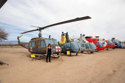 A woman stands beside a row of vintage helicopters in a desert setting, showcasing various military and rescue models.
