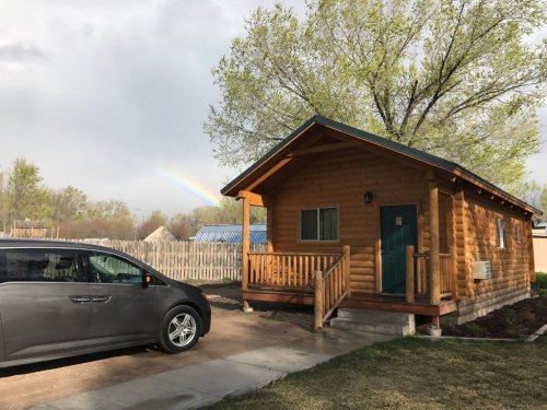 A wooden cabin with a green door and a car parked outside, set against a backdrop of trees and a faint rainbow.