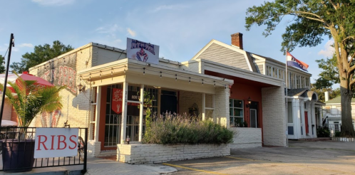 A charming restaurant exterior with a sign reading "RIBS," featuring outdoor seating and a flag in the background.