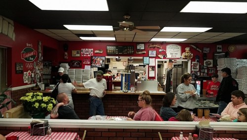 A busy pizzeria interior with customers dining, staff at the counter, and red decor featuring pizza-themed memorabilia.