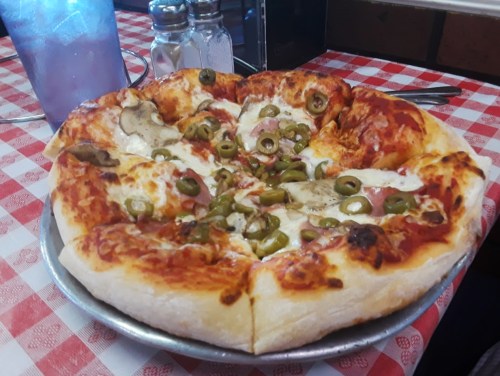 A freshly baked pizza topped with cheese, green olives, and ham, served on a silver platter on a checkered tablecloth.