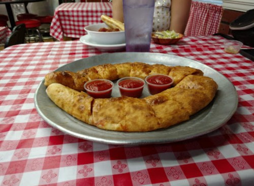 A large, golden-brown calzone on a metal platter, served with three cups of marinara sauce, on a red-checkered tablecloth.