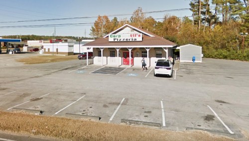 A pizzeria with a brown roof and white exterior, surrounded by a parking lot and trees in the background.
