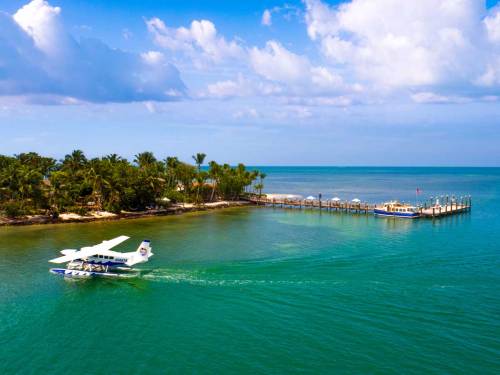 A seaplane glides over turquoise waters near a tropical shoreline and a wooden pier under a blue sky with clouds.