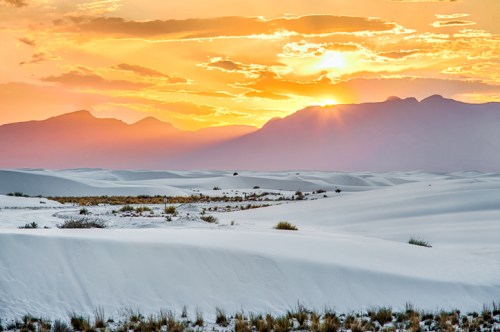 A stunning sunset over white sand dunes, with mountains silhouetted against a colorful sky.
