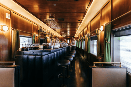 Interior of a vintage train bar with wooden paneling, a long counter, and a bartender preparing drinks.