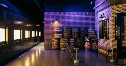 Interior of a dimly lit space featuring brick walls, wooden barrels, and benches, evoking a rustic atmosphere.