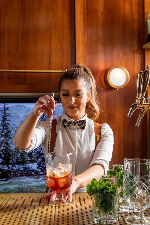 A bartender stirs a cocktail in a glass, surrounded by mint and empty glasses, with a wooden bar backdrop.