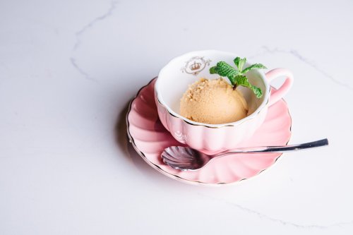 A pink cup with a scoop of ice cream, garnished with a mint leaf, sits on a matching pink plate with a silver spoon.